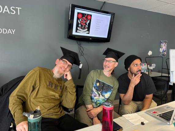 Kenn, Brian and fellow peer Patrick wear black graduation caps with their street clothes sitting at a table.